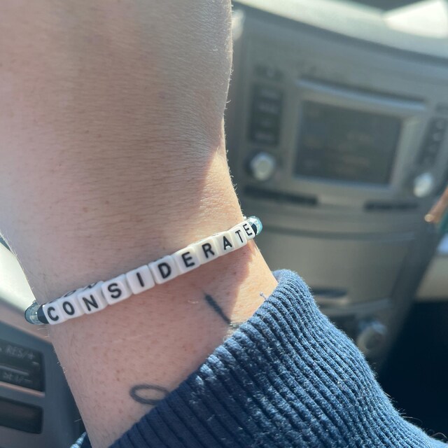 Close-up of a wrist wearing a beaded bracelet that spells 'CONSIDERATE', with the interior of a car visible in the background.