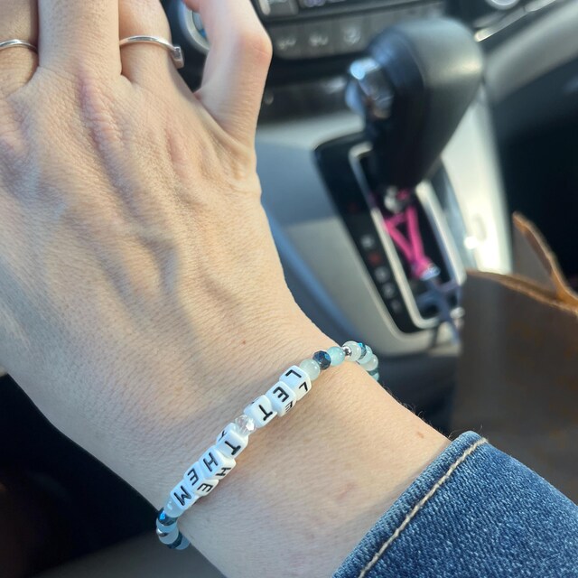 A close-up of a hand wearing a beaded bracelet with the letters 'LET IT BE' on it while in a car.