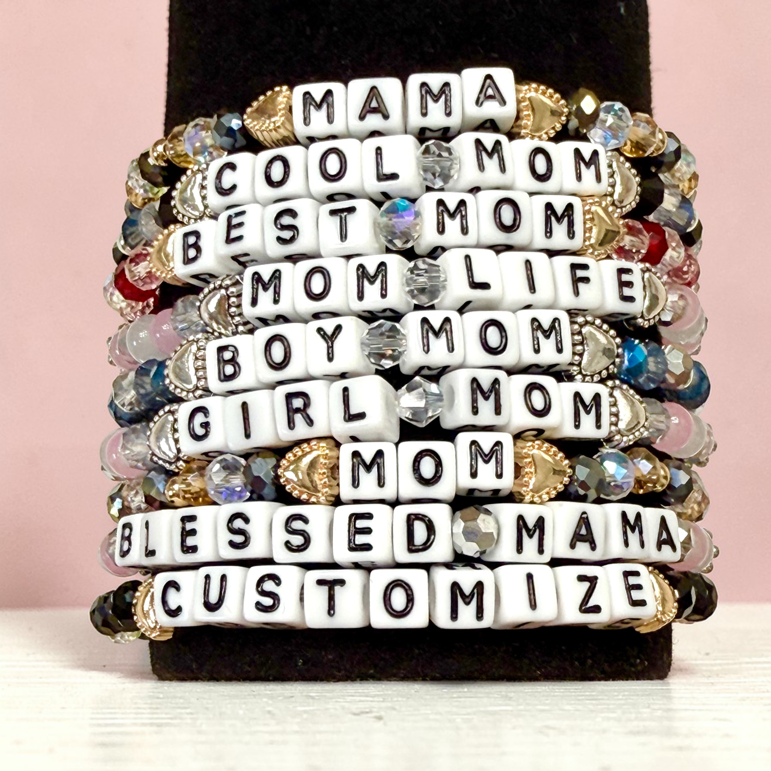 Stack of colorful beaded bracelets featuring word charms like 'Mama', 'Cool Mom', 'Best Mom', 'Mom Life', 'Boy Mom', 'Girl Mom', 'Blessed Mama', and 'Customize' displayed on a stand.